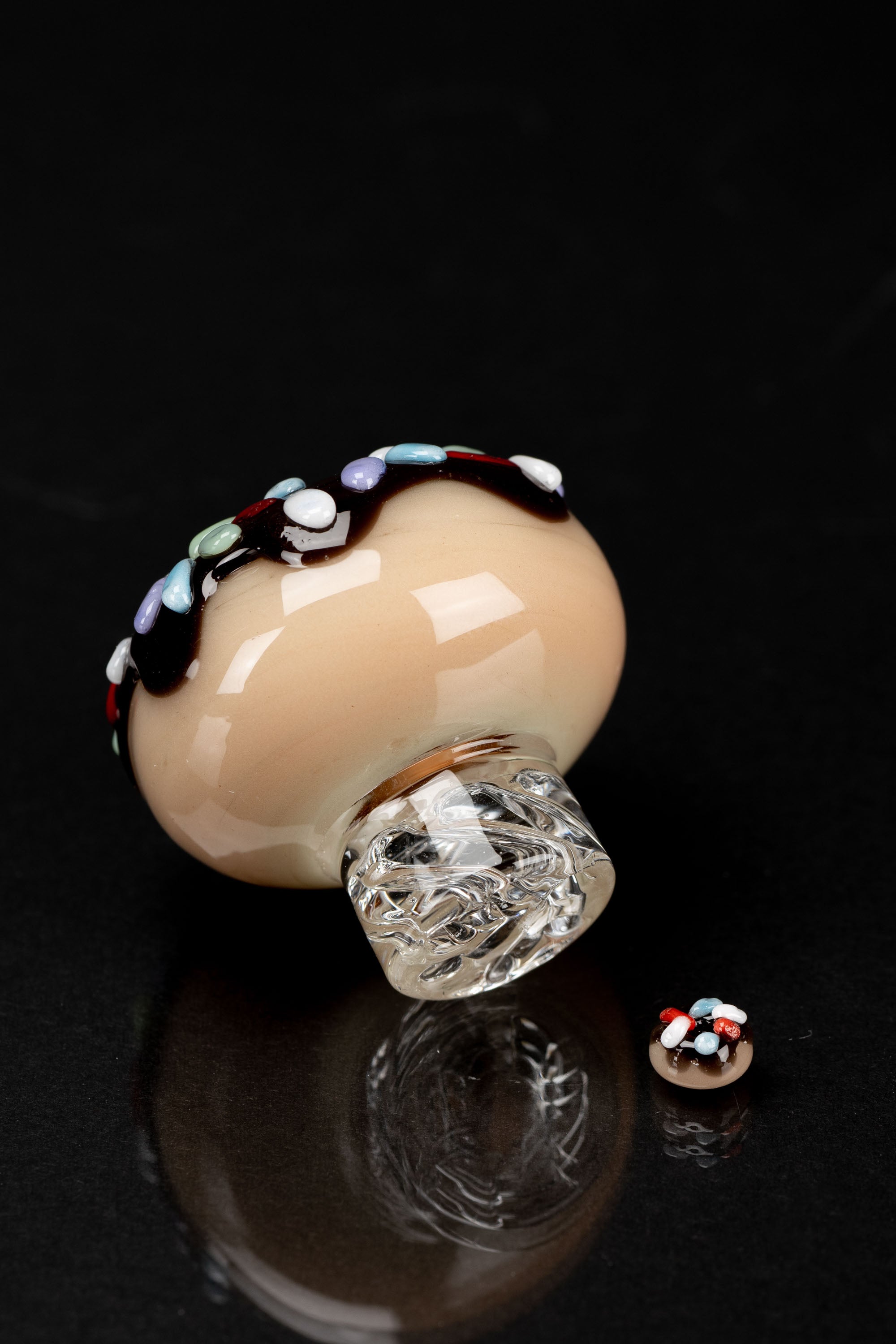 Hand-blown glass mushroom-shaped dab cap with colorful accents and its miniature counterpart, reflected on a dark surface.