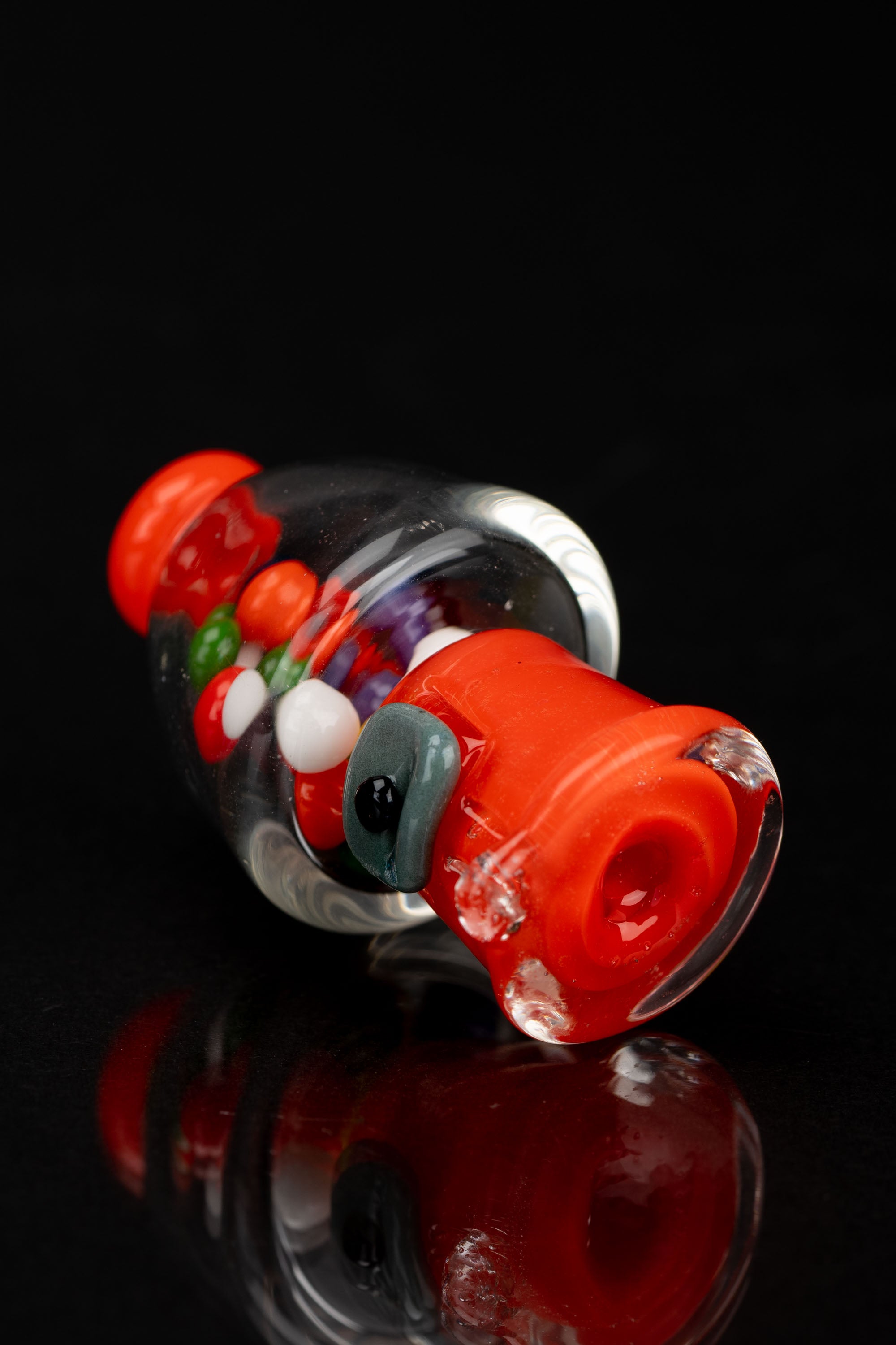 A vibrant red glass pipe with colorful internal patterns and a green mouthpiece, displayed against a black reflective surface.