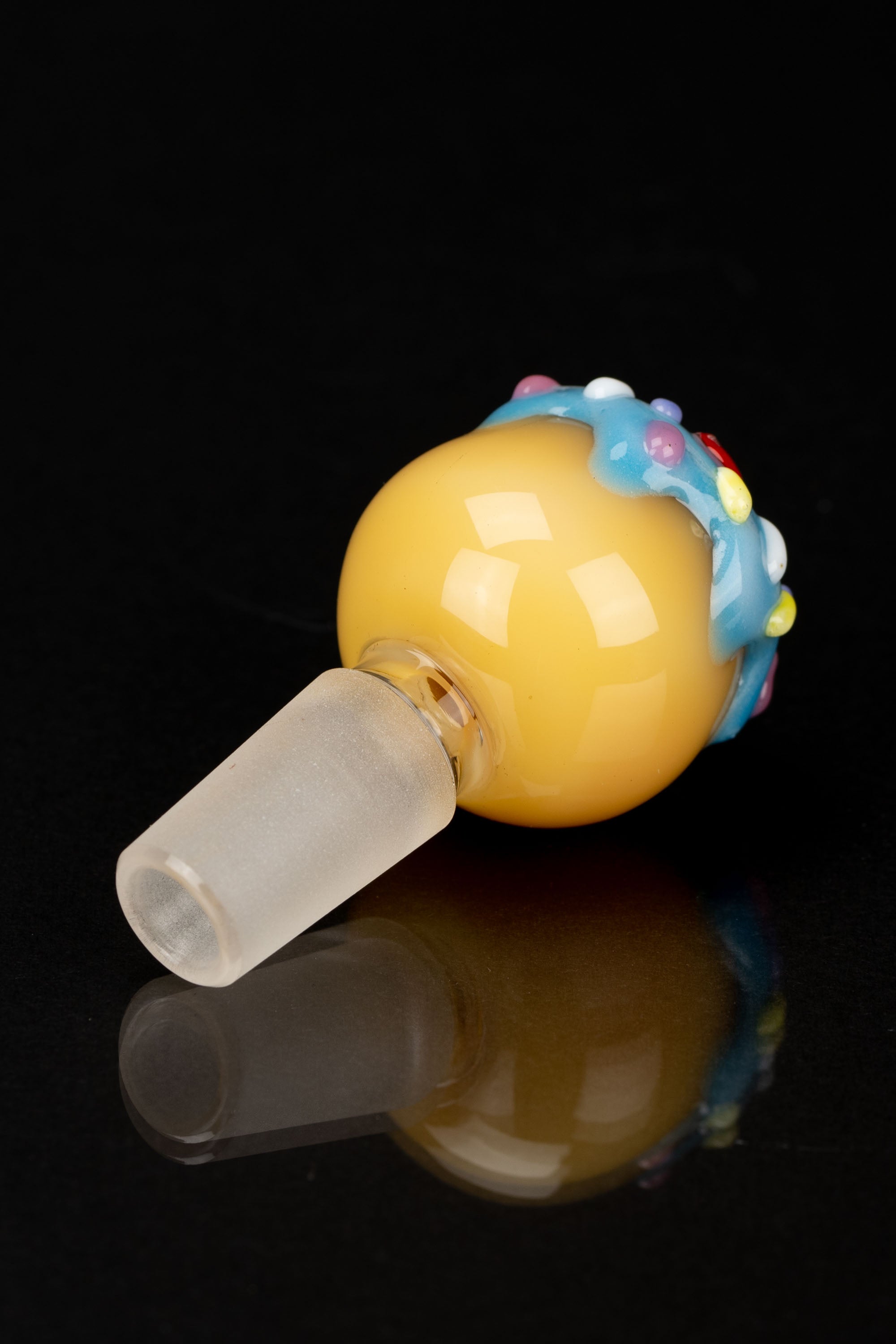 Hand-blown glass cannabis concentrate bowl resembling a donut with colorful sprinkles, reflected on a black surface.