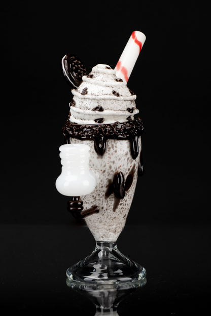 Glass cannabis water pipe shaped like a milkshake with Oreo crumble and chocolate drizzle against a black background.