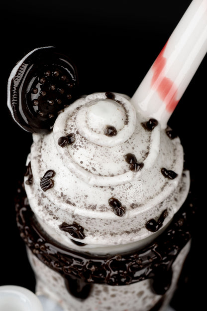 A close-up, top-down view of a chocolate milkshake in a glass, drizzled with chocolate sauce and topped with chocolate chips, alongside a small, dark cannabis accessory.