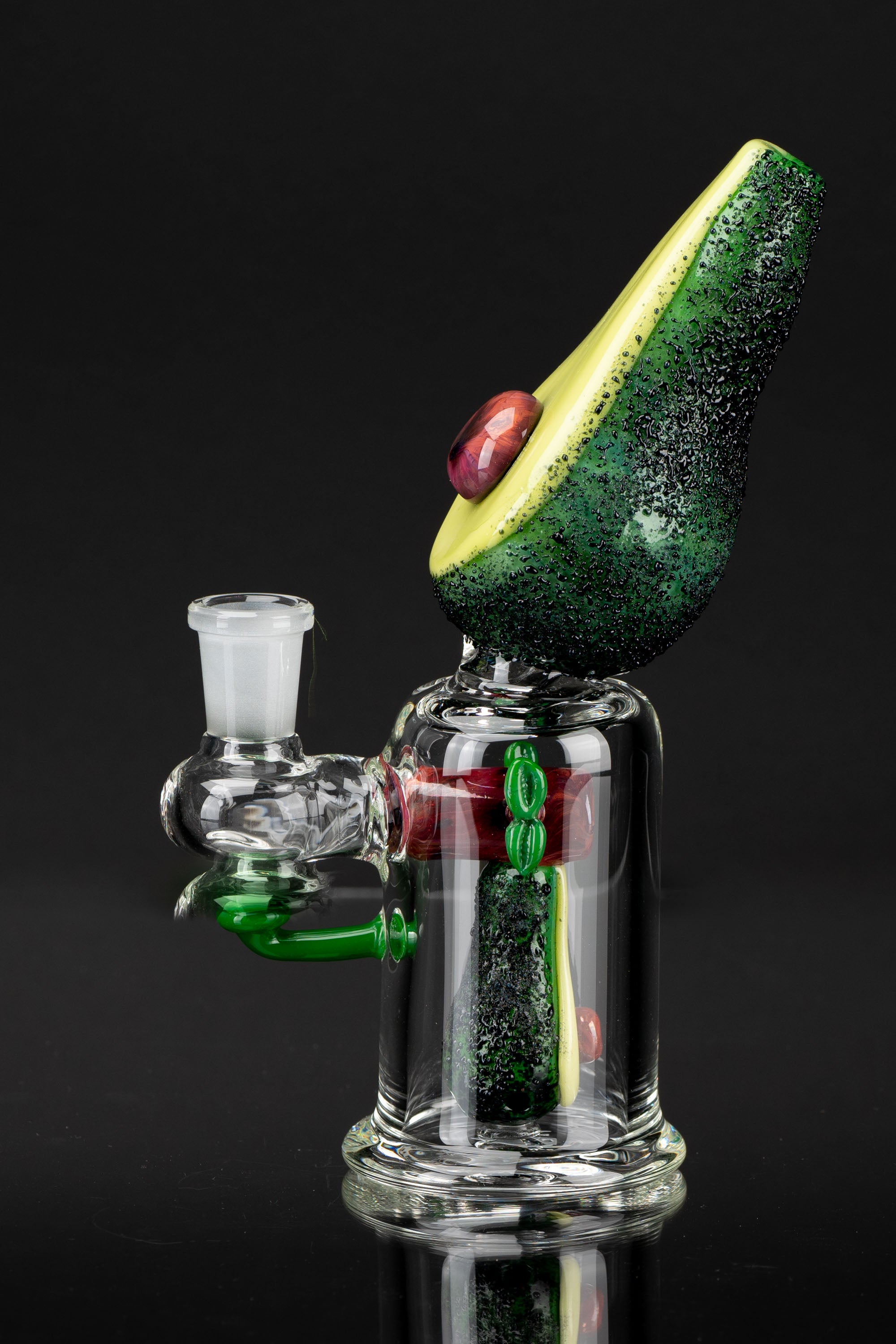 Hand-blown glass water pipe shaped like an avocado, with red and green accents, against a black background.