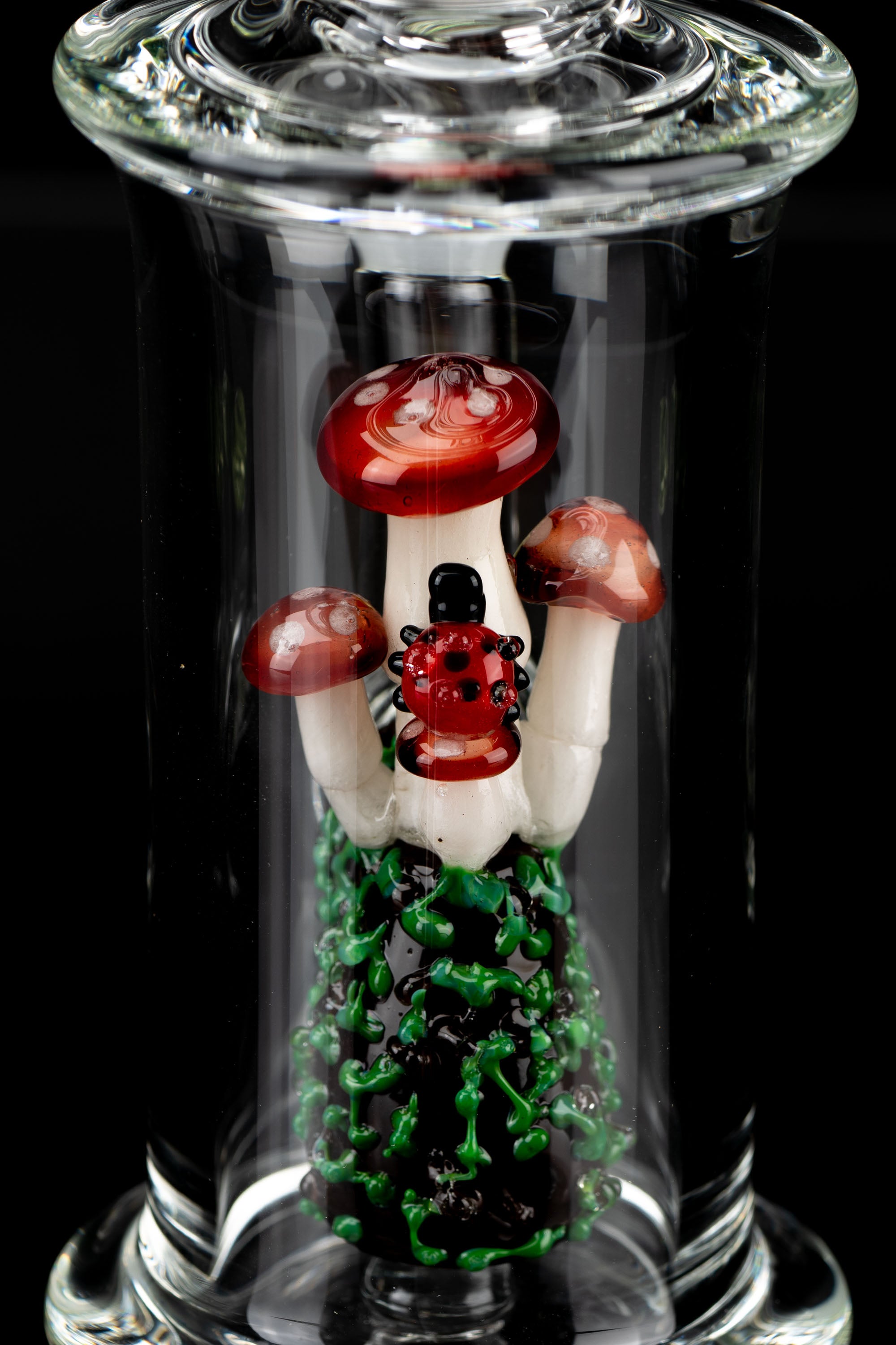 A vibrant glass cannabis pipe sculpted as a cluster of red and white spotted mushrooms with a green base, encased within a clear glass tube.