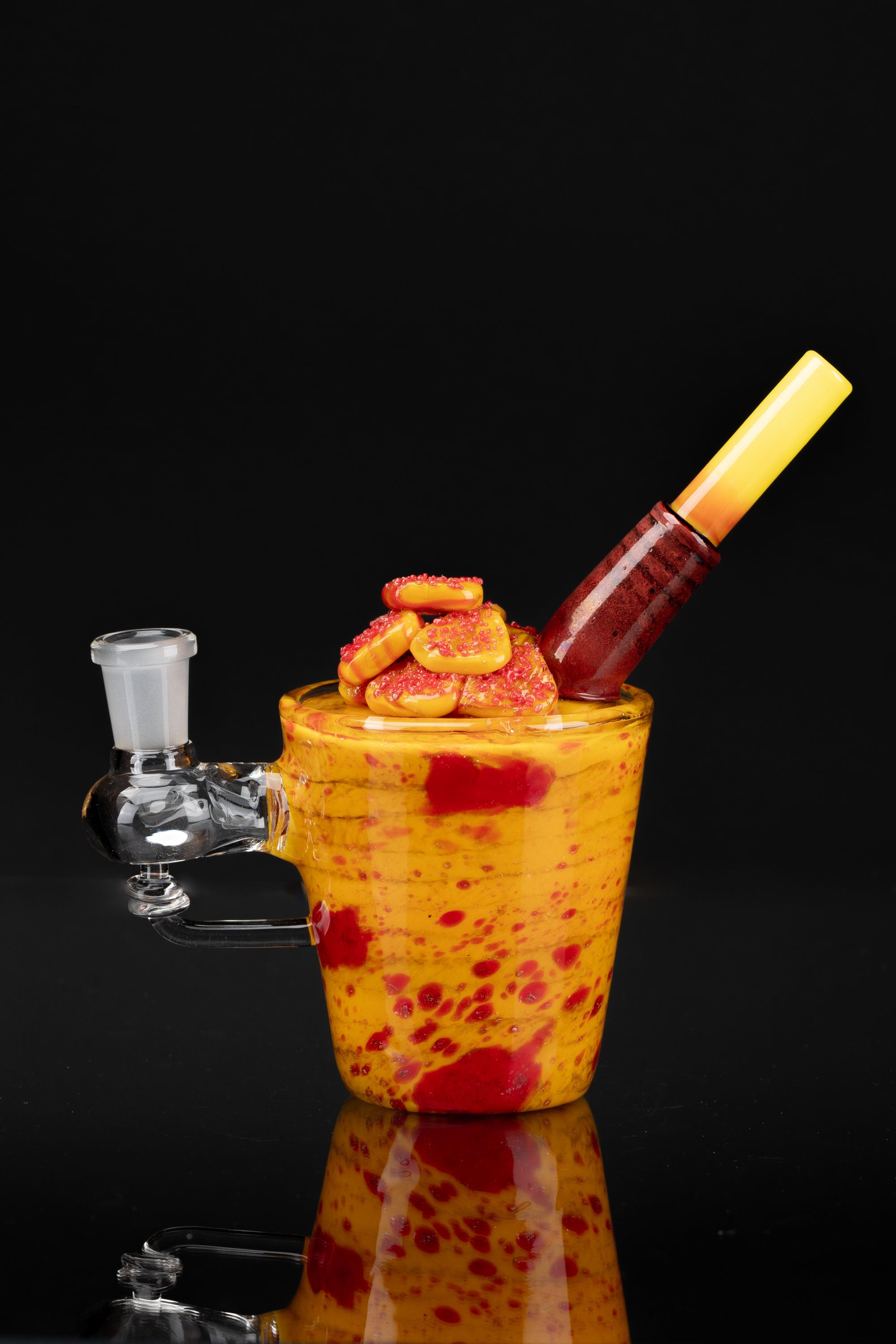 A colorful glass water pipe filled with cannabis candies and topped with a glass smoking tool, reflecting on a dark surface.