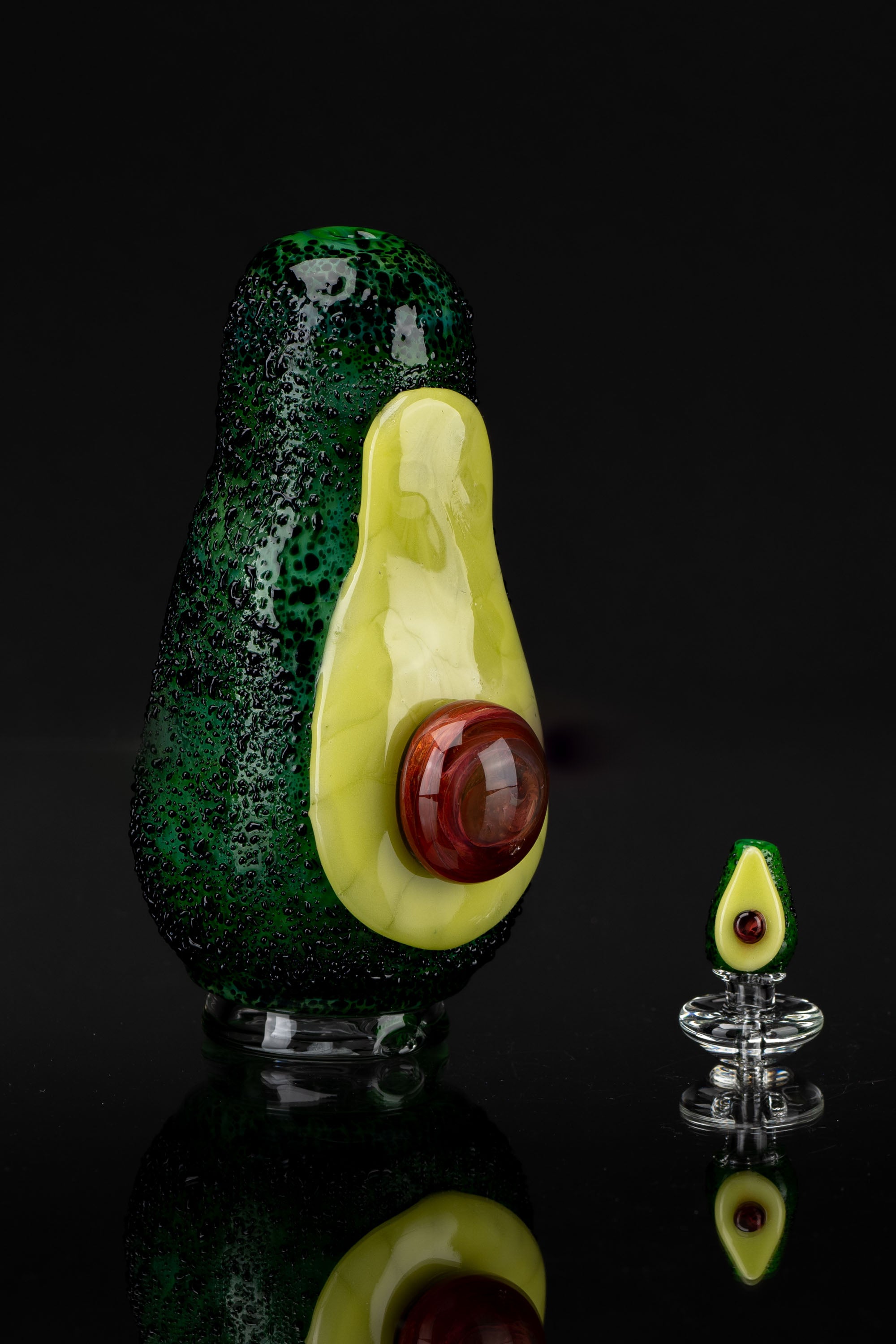 Hand-blown glass avocado-shaped cannabis water pipe with a red ""pit"" mouthpiece, reflected on a black surface."