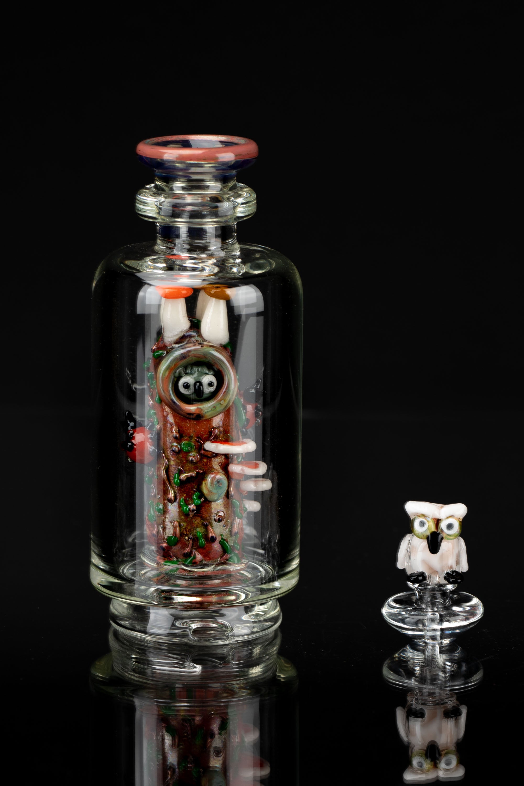 Hand-blown glass bubbler featuring a whimsical, cartoonish monster design with intricate swirling colors, displayed alongside its matching cap, both reflected on a dark surface.