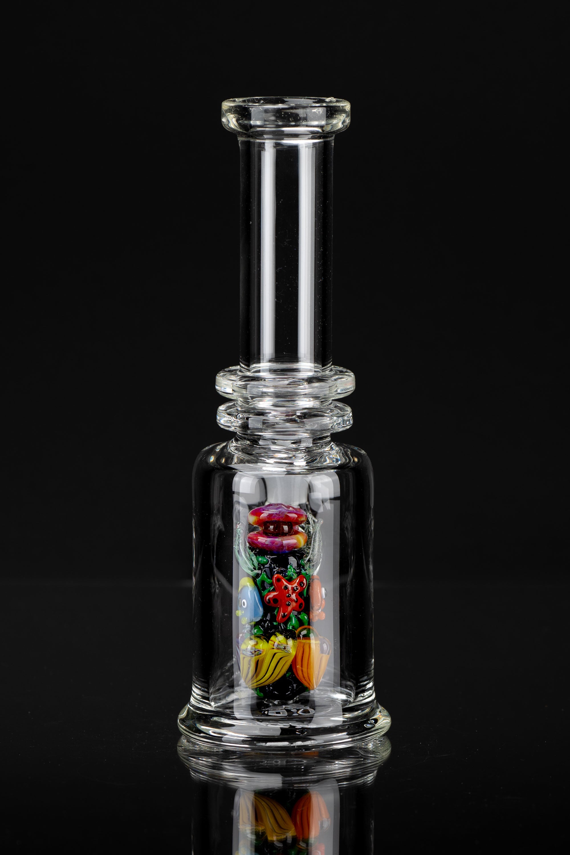 Clear glass bubbler water pipe with colorful glass insects suspended within, reflecting on a dark surface.
