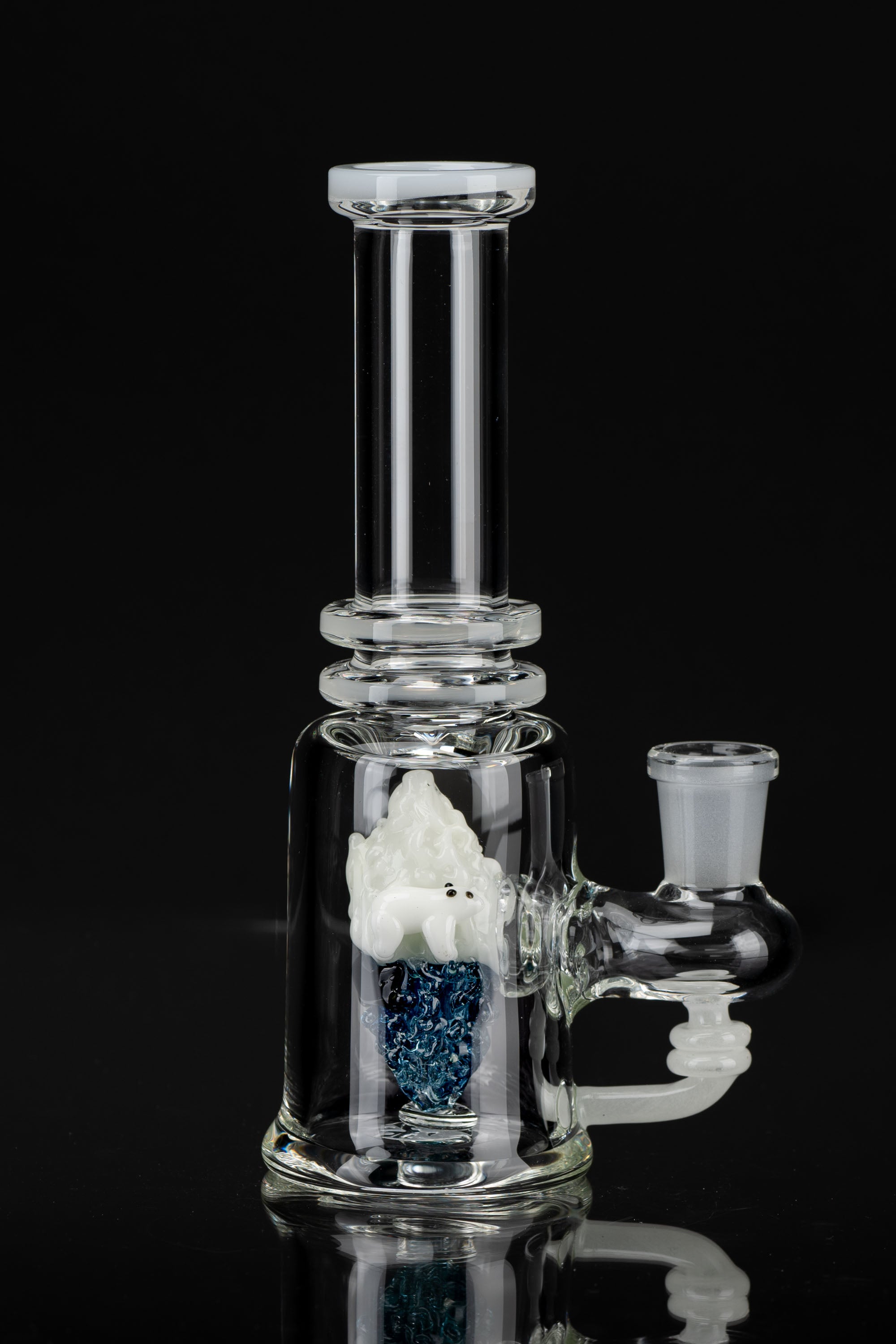 Clear glass water pipe (bong) with a sculpted gnome figure inside a blue glitter chamber, reflecting on a black surface.