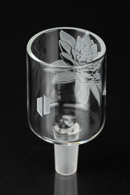 Clear glass cannabis concentrate container (dab dish) with etched floral design and manufacturer's logo, isolated on black.