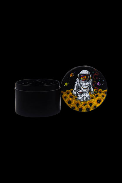 Black grinder with a lid displaying an astronaut amongst sunflowers and planets, cannabis-themed art.