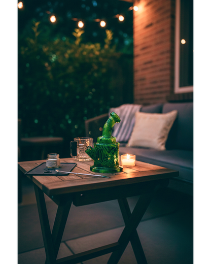 Evening view of an Eyce ORAFLEX Rig in swirled green glass on a wooden patio table beside a lit candle and jar.