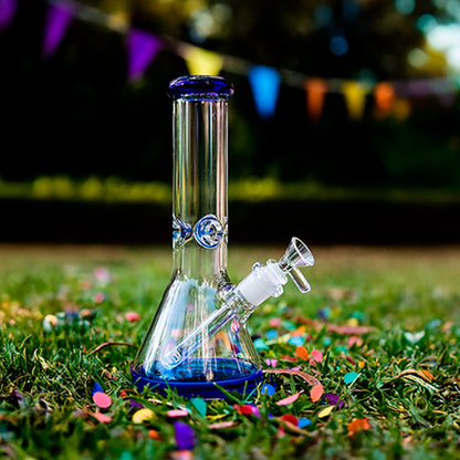 Front view of the Smoke Cartel 9mm Big Base Beaker w/ Color Accents Bong - 12 in. resting on grass with blue-accented glass and colorful confetti.