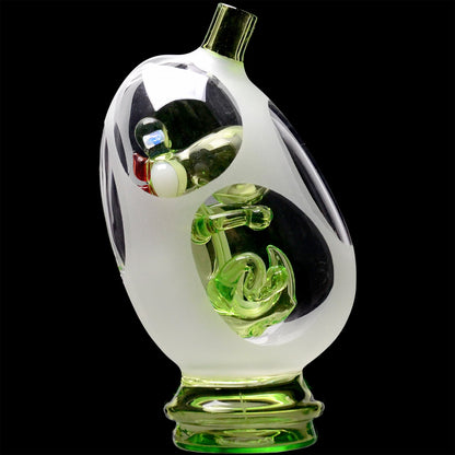 Close-up angled view of the Glassic Frosted Dino Egg Puffco Peak Glass Attachment showing frosted outer glass and a green sculpted dino encased against a black background