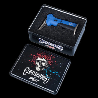 Angled top-down view of a blue Grateful Dead Alternator Torch nestled in black foam inside a metal tin with matching skull-and-roses lid.