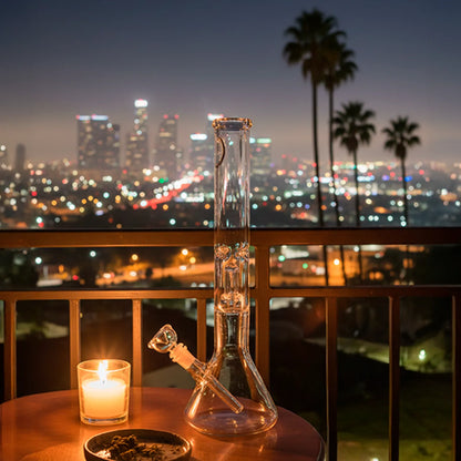 Night view of a clear LA Pipes 5mm Thick Beaker Bong with Showerhead Perc standing on a balcony table beside a lit candle.