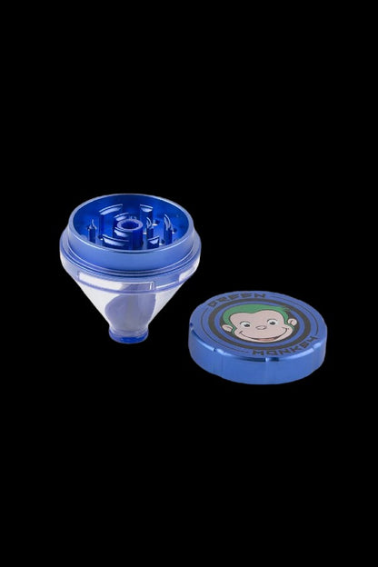 Angled view of a disassembled Green Monkey Patas Grinder in glossy black-blue plastic on a black background, showing the chamber with teeth and the lid with a smiling monkey logo