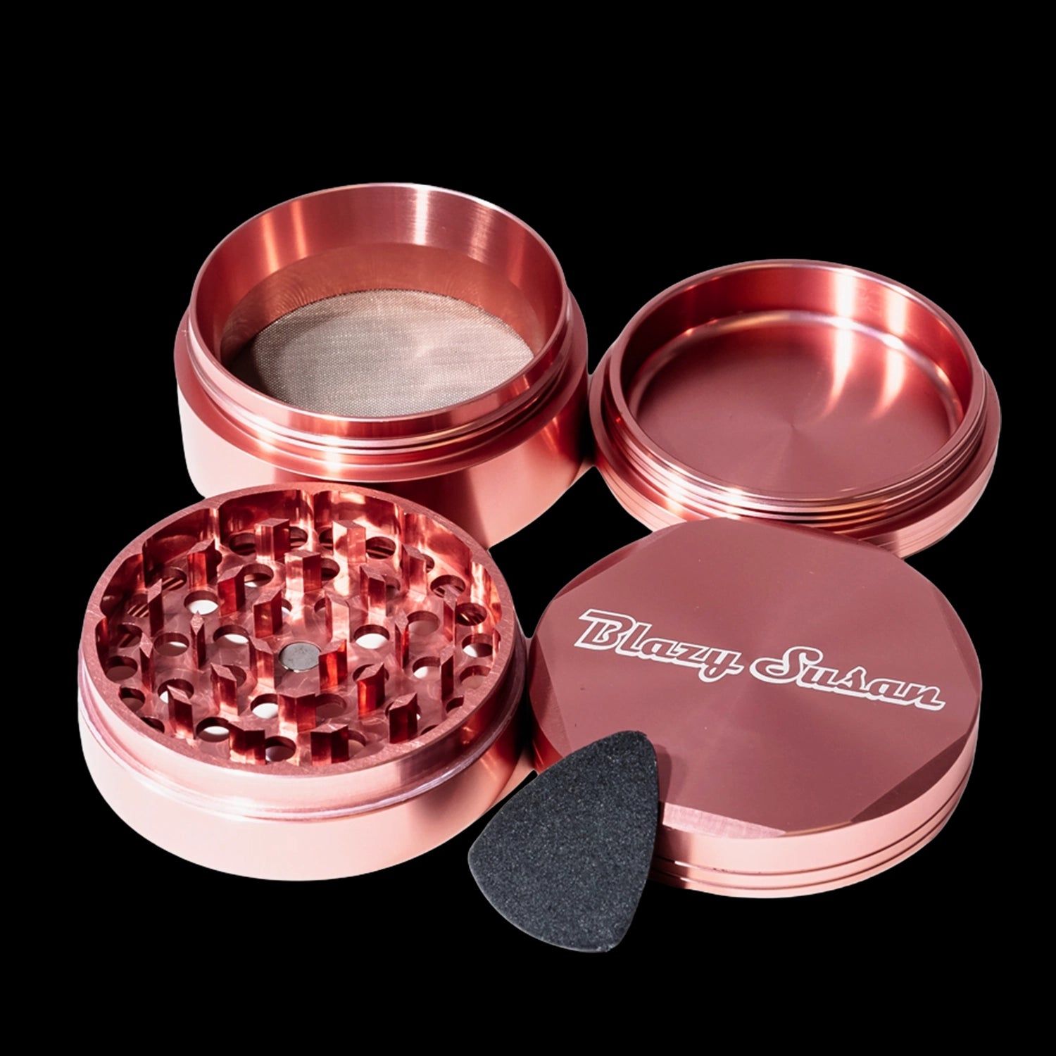 Angled close-up of a disassembled rose-gold Blazy Susan 4-pc 3 Chamber Grinder showing the textured grinding plate, mesh screen chamber, lid with logo, and a black scraper.