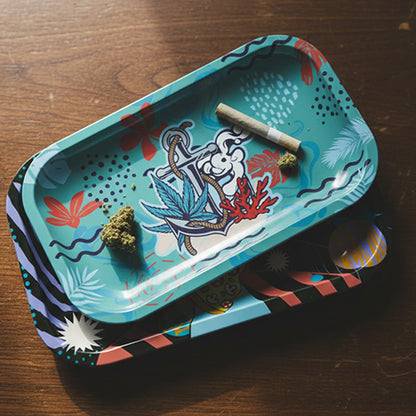 Top-down view of the Atomik Rolling Tray - Medium on a wooden surface showing its teal tropical anchor illustration, scattered buds, and a rolled joint.