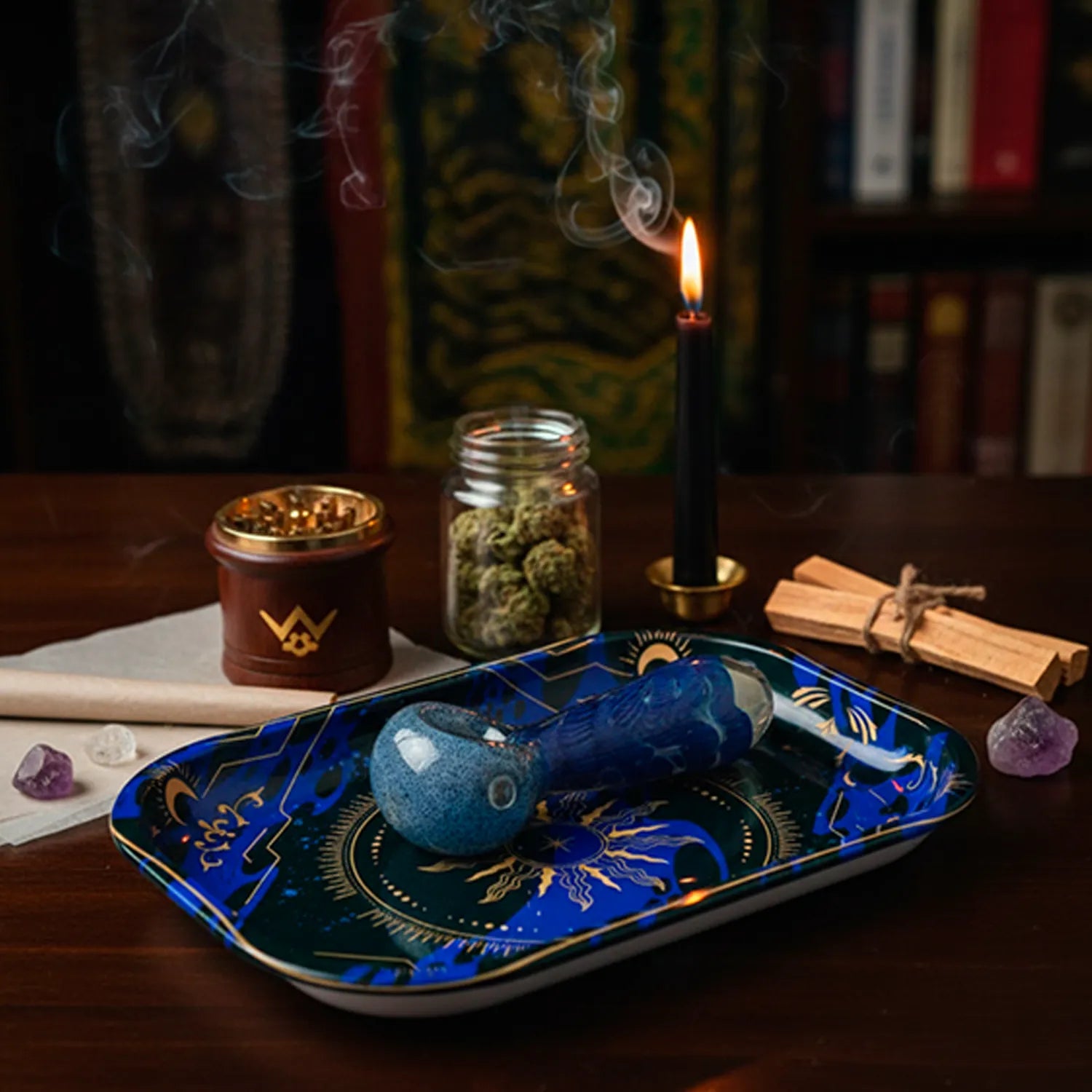 Angled close-up of the Famous Design - Astral Power Rolling Tray Small showing a blue glass pipe resting on its celestial patterned surface with a lit black candle and jar of buds in the background.