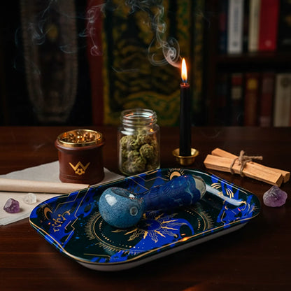Angled close-up of the Famous Design - Astral Power Rolling Tray Small showing a blue glass pipe resting on its celestial patterned surface with a lit black candle and jar of buds in the background.
