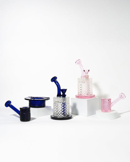 Blue and pink cannabis accessories – including dab rigs, bongs, and a dish – displayed on white pedestals against a plain white background.