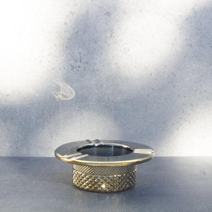 Gold cannabis ashtray with a textured, patterned base, set against a muted, textured wall.