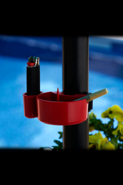 Red cannabis accessory holder attached to a black pole, containing a lighter and rolled joint, with blurred green foliage and a blue background.
