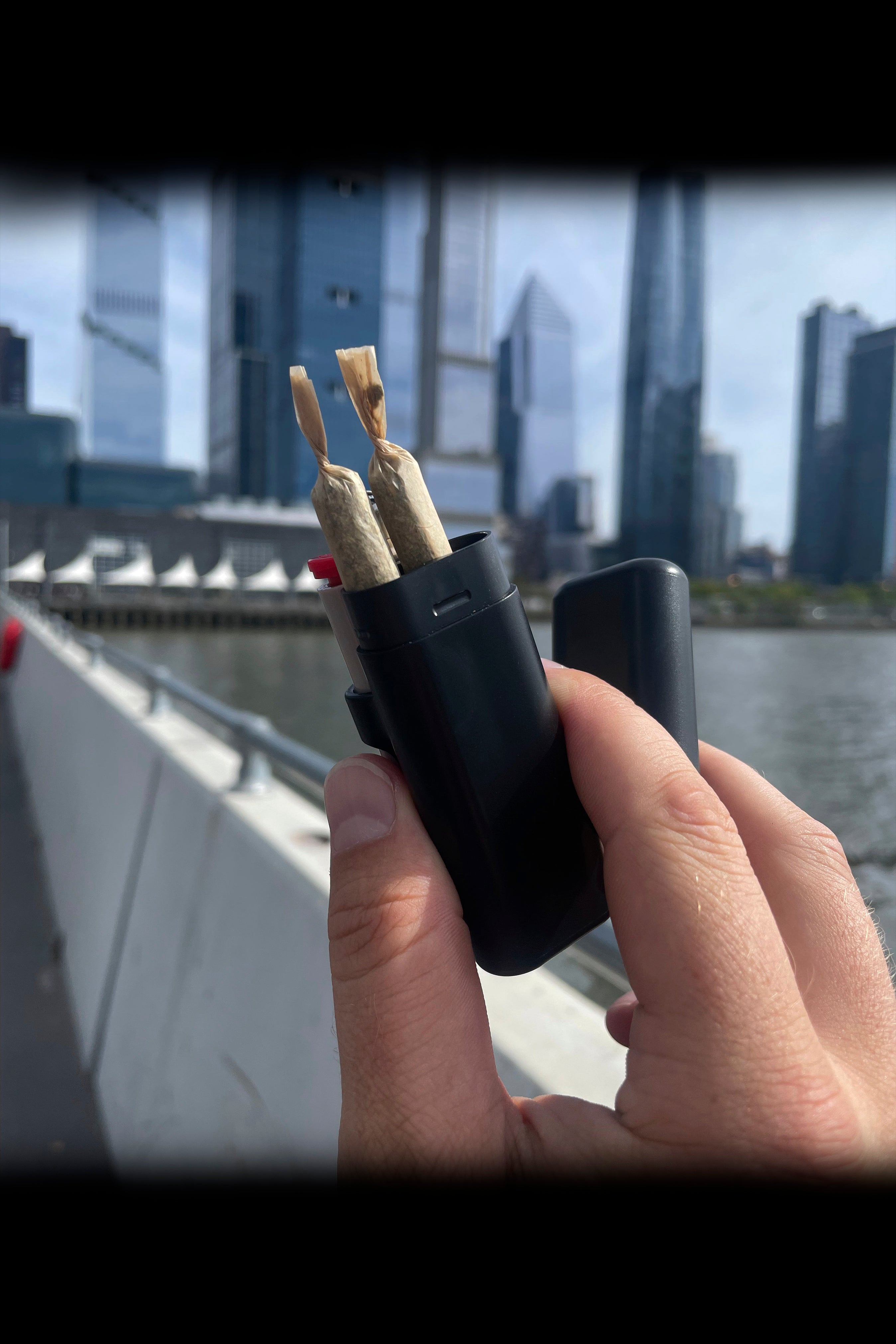 Hand holding a black case containing two rolled cannabis cigarettes with a cityscape blurred in the background.