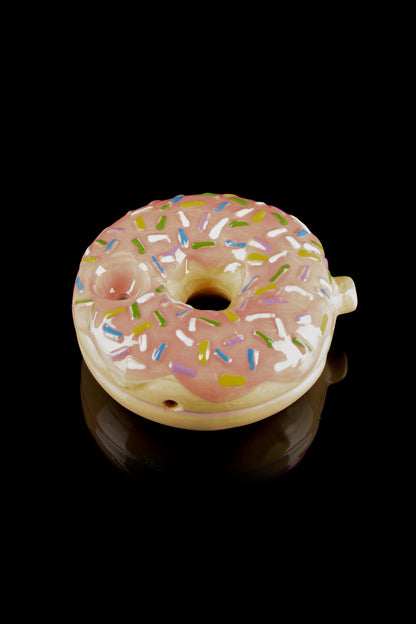 Pink glazed donut-shaped glass piece with colorful sprinkles, reflecting on a dark surface."