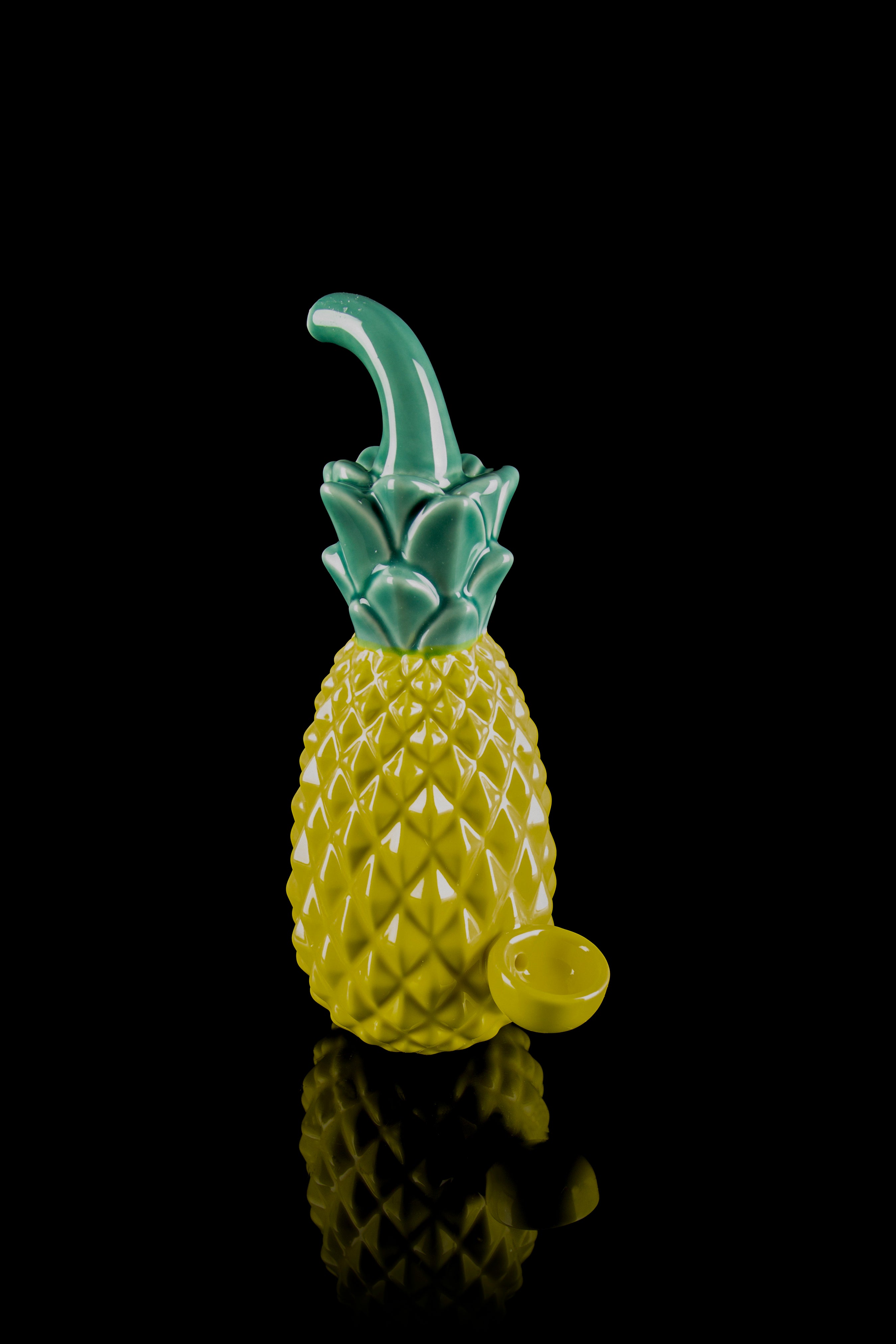 A glass pineapple-shaped water pipe with a green stem and base, reflected on a black surface.