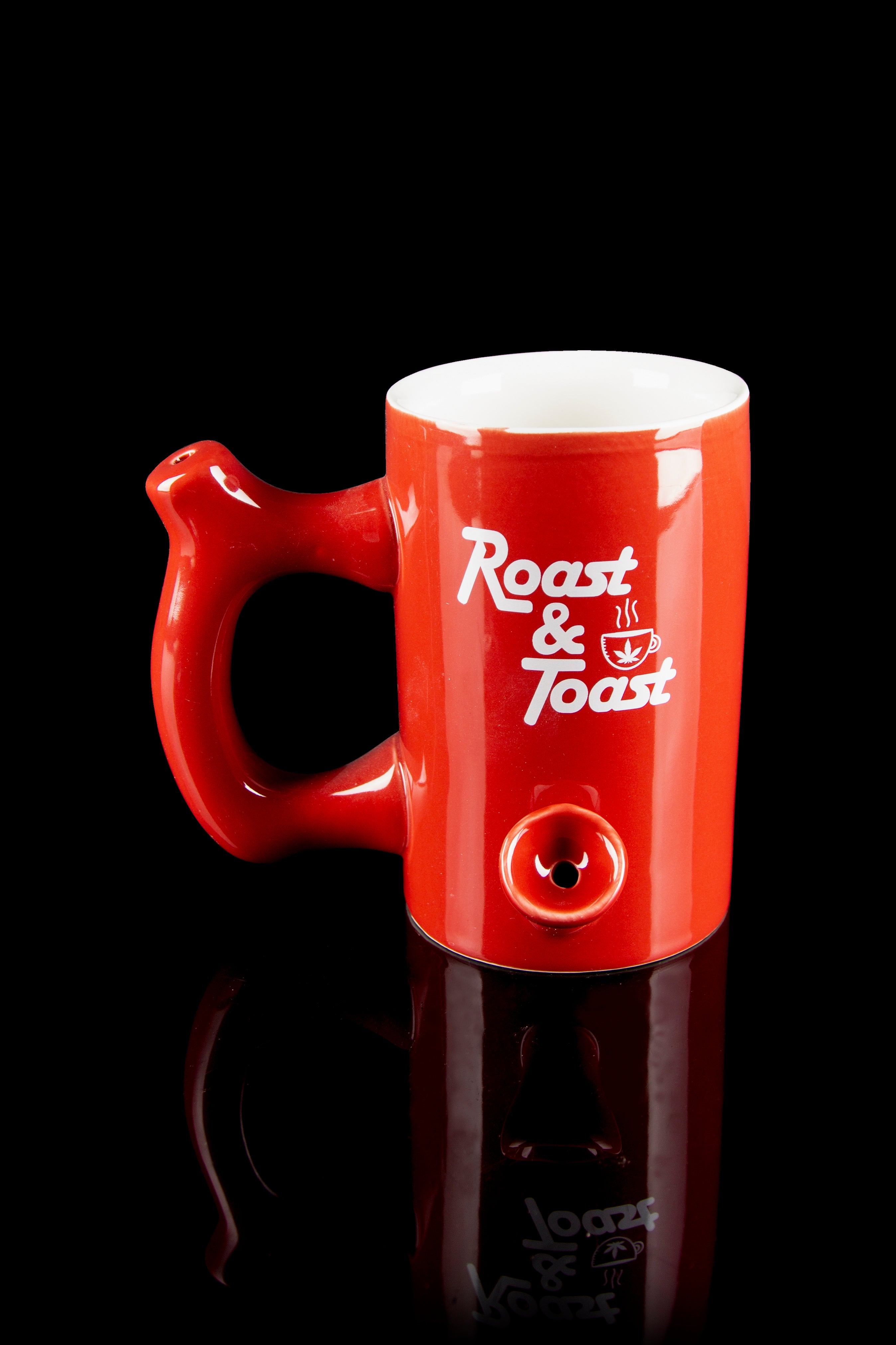 Red ceramic mug with a built-in pipe, featuring "Roast & Toast" lettering and a cannabis leaf design, displayed on a reflective surface.