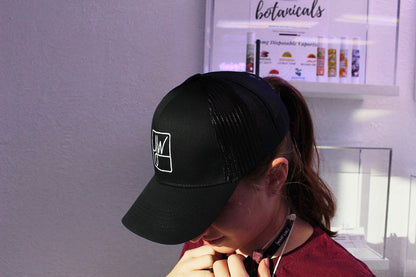 A person wearing a black trucker hat with a white logo, standing in front of a display of cannabis vaporizers.