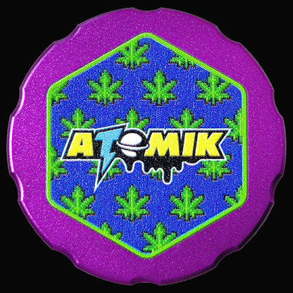 Purple grinder with a blue and green hexagonal cannabis leaf design and the brand name "ATOMIK" in yellow and blue.