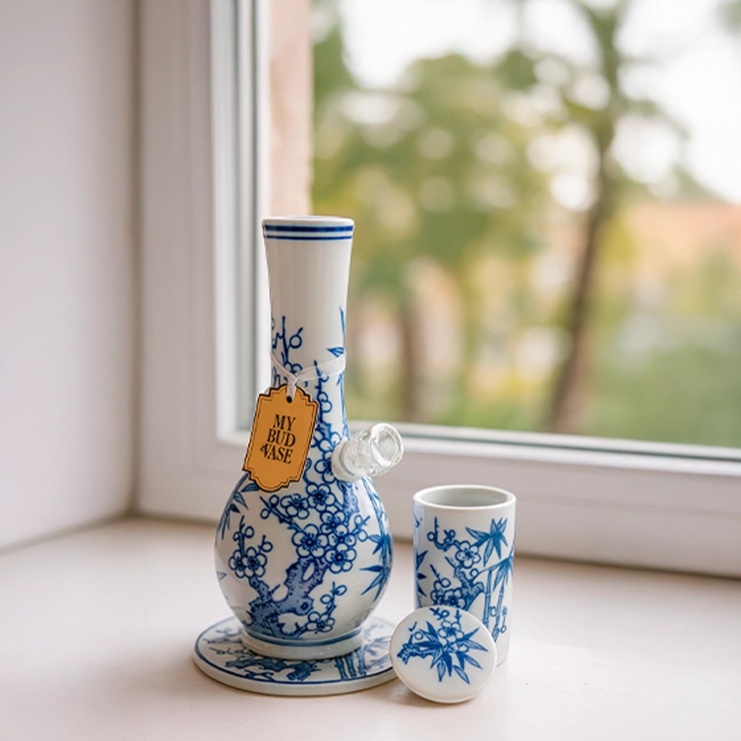 Three-quarter view of the blue-and-white porcelain My Bud Vase "Luck" Porcelain Vase Bong sitting on a windowsill with matching cup and lid.