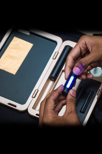 Hands holding a lit, glowing cannabis vaporizer pen over an open, wood-toned cannabis accessory case with rolled joints and a grinder inside.
