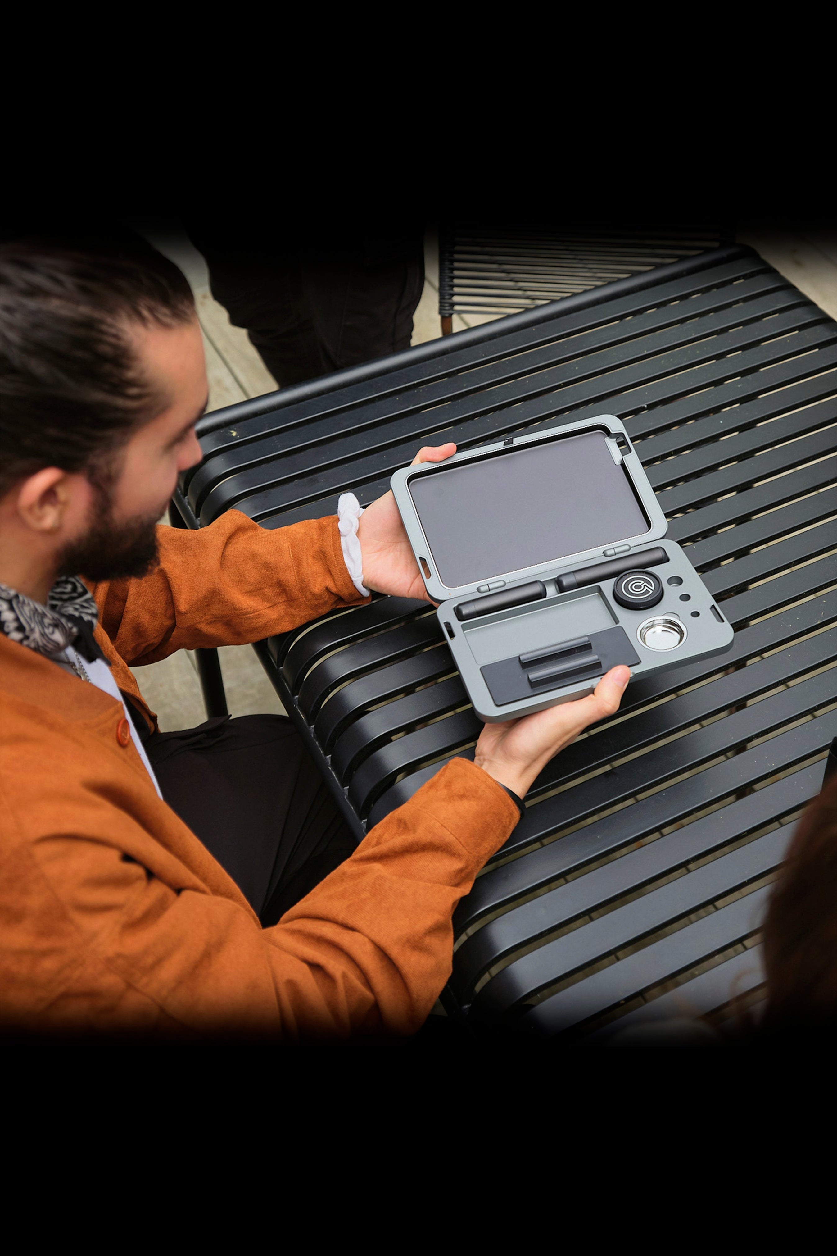 Man holding a grey cannabis accessory case open on a slatted surface, revealing organized compartments.