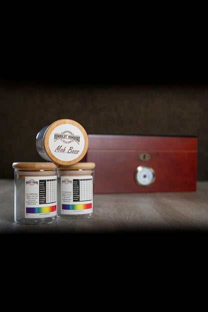 A still life of cannabis accessories including a wooden humidor labeled “Mob Boss”, two glass jars with colorful cannabis strain information, and a small metallic sculpture, all on a reflective black surface.