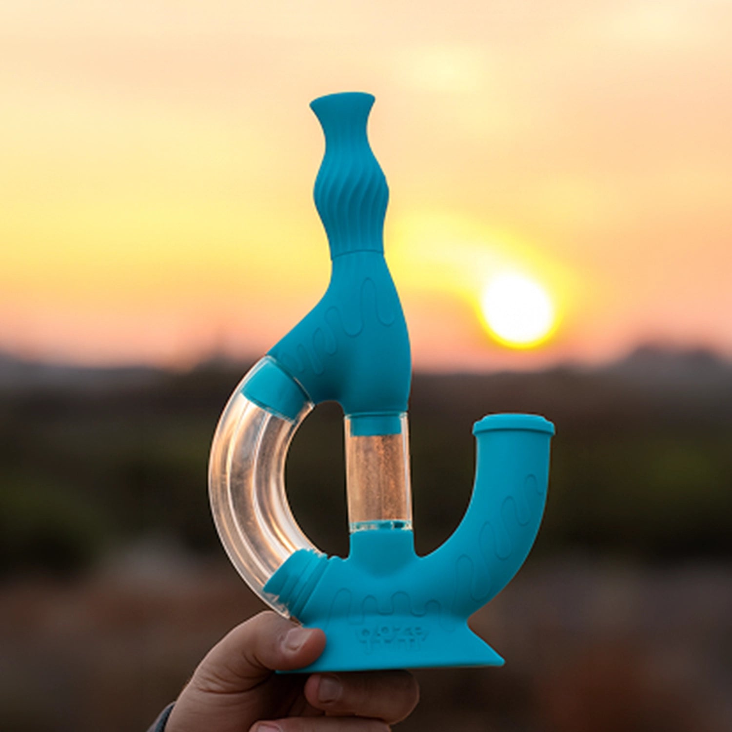Handheld against a glowing sunset, the blue Ooze Echo Silicone Bong shows its curved clear tube and textured silicone body from a side angle.