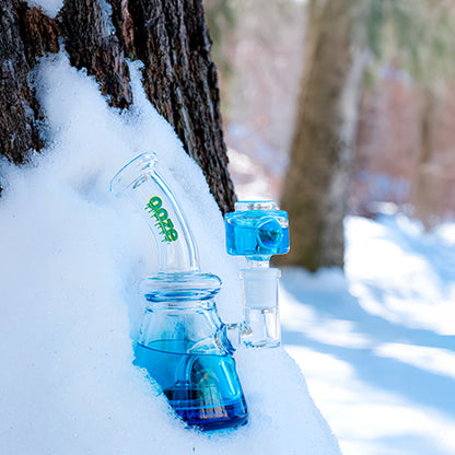 Angled close-up of the Ooze Glyco Glycerin Chilled Bong nestled in snow against a tree trunk, showing its blue water chamber and green logo.