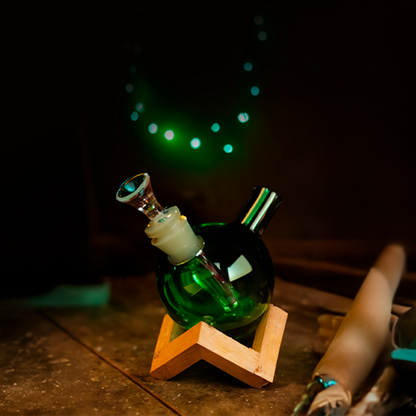 Three-quarter view of the teal Art of Smoke Orb Bubbler resting in a wooden star stand against a dark background with circular green bokeh lights.
