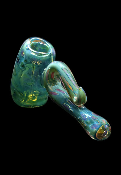 Angled close-up of the Smoke Cartel Gold Fume Bubble Trap Hammer Bubbler - 6 in., showing its iridescent green-gold glass with a bulbous chamber and curved handle against a black background