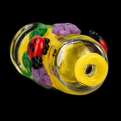 Close-up angled view of the Empire Glassworks Puffco Pivot Glass Mouthpiece - Sunshine Garden showing a yellow glass mouthpiece adorned with purple flowers and a red ladybug on a black background