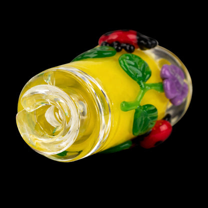 Close-up angled view of the Empire Glassworks Puffco Pivot Glass Mouthpiece - Sunshine Garden showing a yellow glass body with raised green leaves, a purple flower and a red ladybug on a black background