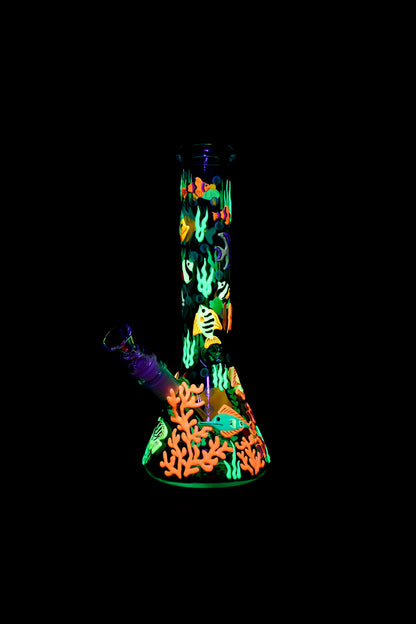 Glowing glass water pipe decorated with vibrant coral reef and fish designs against a black background.
