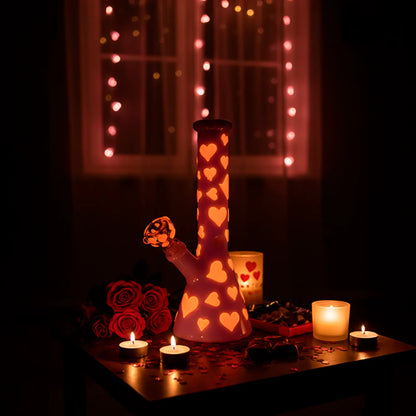 Center-framed Valentines Day Heart Glow In The Dark Beaker glowing with heart cutouts on a candlelit table against pink string lights