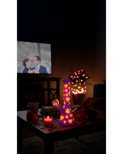 Nighttime front view of the Valentines Day Heart Glow In The Dark Beaker glowing purple with heart cutouts on a coffee table.