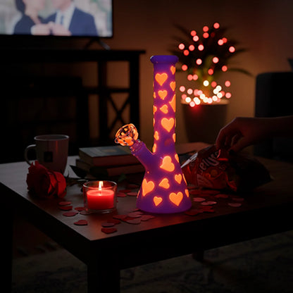 A dimly lit front view of the Valentines Day Heart Glow In The Dark Beaker glowing purple with orange heart cutouts on a coffee table amid rose petals and a lit candle