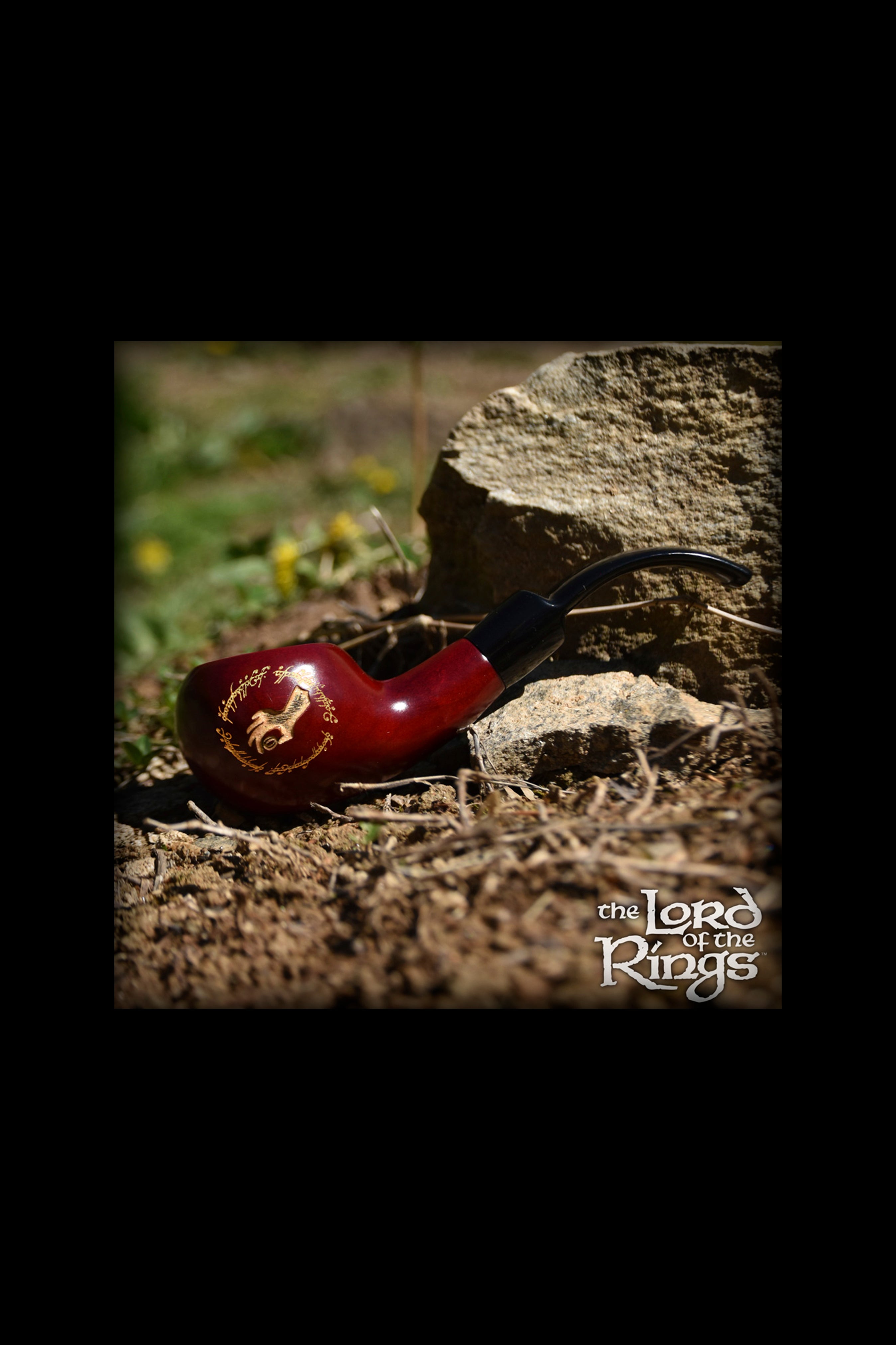 A red glass pipe with intricate gold detailing rests on a weathered tree stump amidst green grass and brown earth, evoking a natural, earthy aesthetic alongside “The Lord of the Rings” logo.