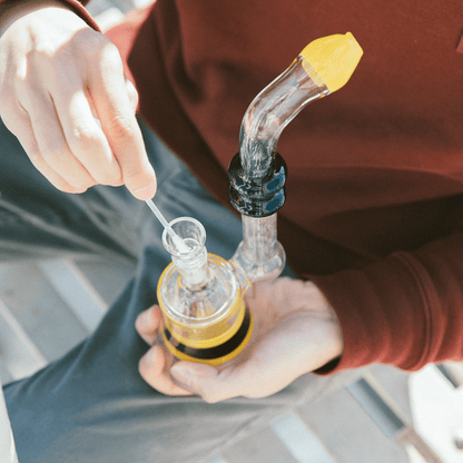 Top-down view of hands holding a glass rig while using Hemper Tech Cotton Buds 200ct to clean the bowl against a blurred outdoor background