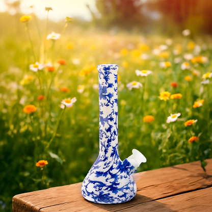 Close-up of a blue-and-white floral patterned BONGS USA Floral Silicone Bong standing upright on a wooden bench with a sunlit wildflower meadow blurred behind it